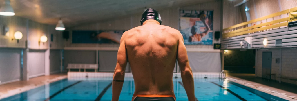 In einer gut beleuchteten Schwimmhalle steht ein Schwimmer ohne Hemd und Kappe am Beckenrand und blickt entschlossen ins Wasser.